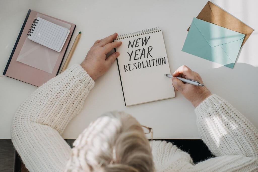 Healthy New Years resolutions for older adults. Senior mature old woman in white sweater writing handwritten text New Years resolutions in open notepad on the table. Planning and setting goals